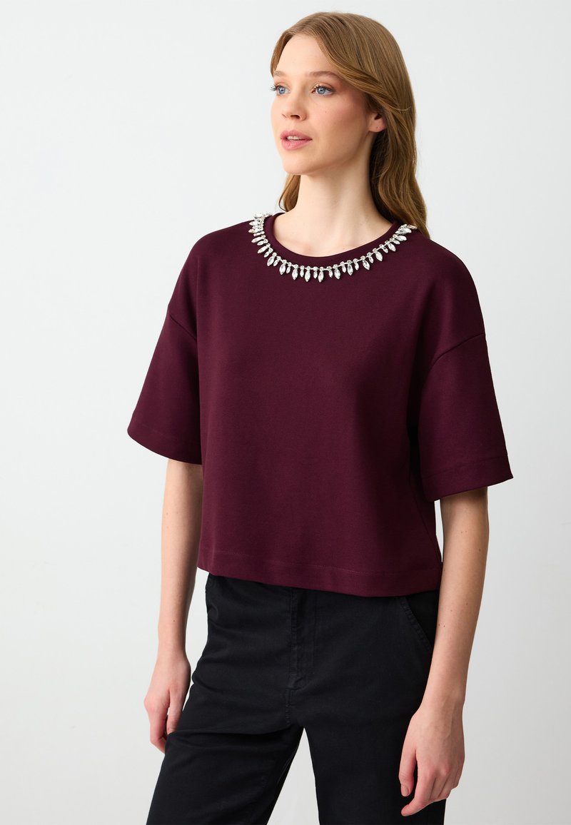 Bordeaux geknipte t-shirt met korte mouwen en een decoratieve kraalrand. Zacht materiaal met een casual pasvorm, gecombineerd met zwarte broek.
