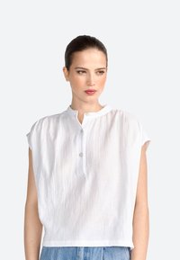 Blouse blanche à manches courtes et texturée, avec col montant et fermeture à un bouton au cou. Conçu avec une coupe décontractée.