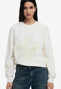 Light beige sweatshirt with a textured flower design, featuring ribbed shoulder details and a rounded neckline, paired with dark jeans.