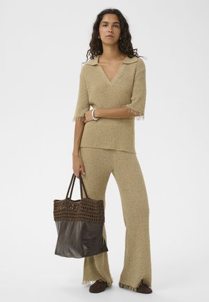 Naine beežis kootud ülaosa ja pükstega, kes hoiab suurt pruuni käekotti, seisab ühevärvilise heleda tausta ees.