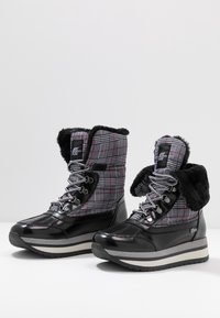 Paire de bottes d'hiver noires et à carreaux avec semelles en caoutchouc épaisses, devant à lacets et doublure en fourrure noire, présentées sur une surface blanche.