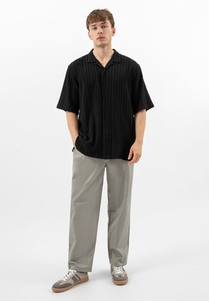 Mladý muž stojící v černé krátký rukáv košili na zapínání, světle šedých kalhotách a šedých teniskách s bílými tkaničkami.