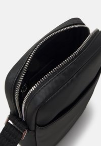 Borsa sintetica nera con superficie texturizzata, chiusura con zip e tracolla regolabile. L'interno presenta una fodera liscia e uno slot per carte.