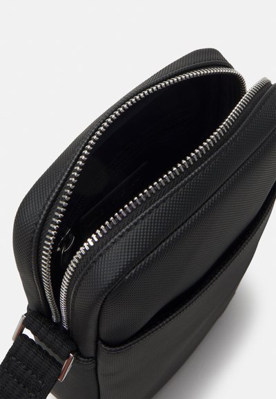 Sac en synthétique noir avec une surface texturée, fermeture éclair et bandoulière ajustable. L'intérieur présente une doublure lisse et un emplacement pour cartes.