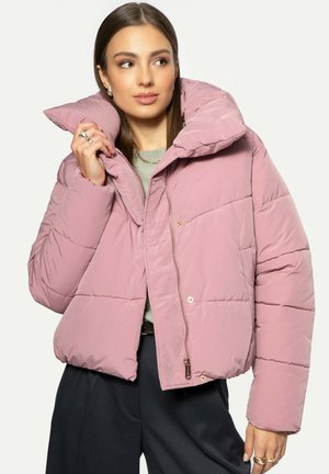 Mladá žena upravující růžovou vatovanou bundu s vysokým límcem, má na sobě světle zelený top a tmavé kalhoty na jednobarevném pozadí.