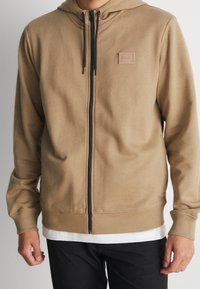 Hellbrauner Kapuzen-Sweatshirt mit glatter Textur, ausgestattet mit einem schwarzen Reißverschluss und einem kleinen Markenpatch auf der linken Brust.