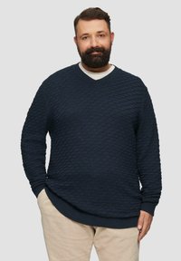 Granatowy sweter z dzianiny w wzór diamentowy, z dekoltem w kształcie litery V i teksturowanym materiałem. Połączony z beżowymi spodniami. Casualowy krój.