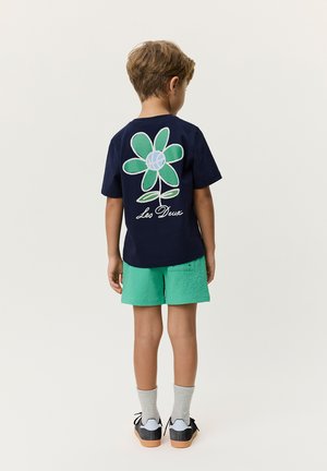 Gutt som står med ryggen til, iført marineblå t-skjorte med grønn blomst og teksten "Les Deux", grønne shorts, grå sokker og svarte joggesko.
