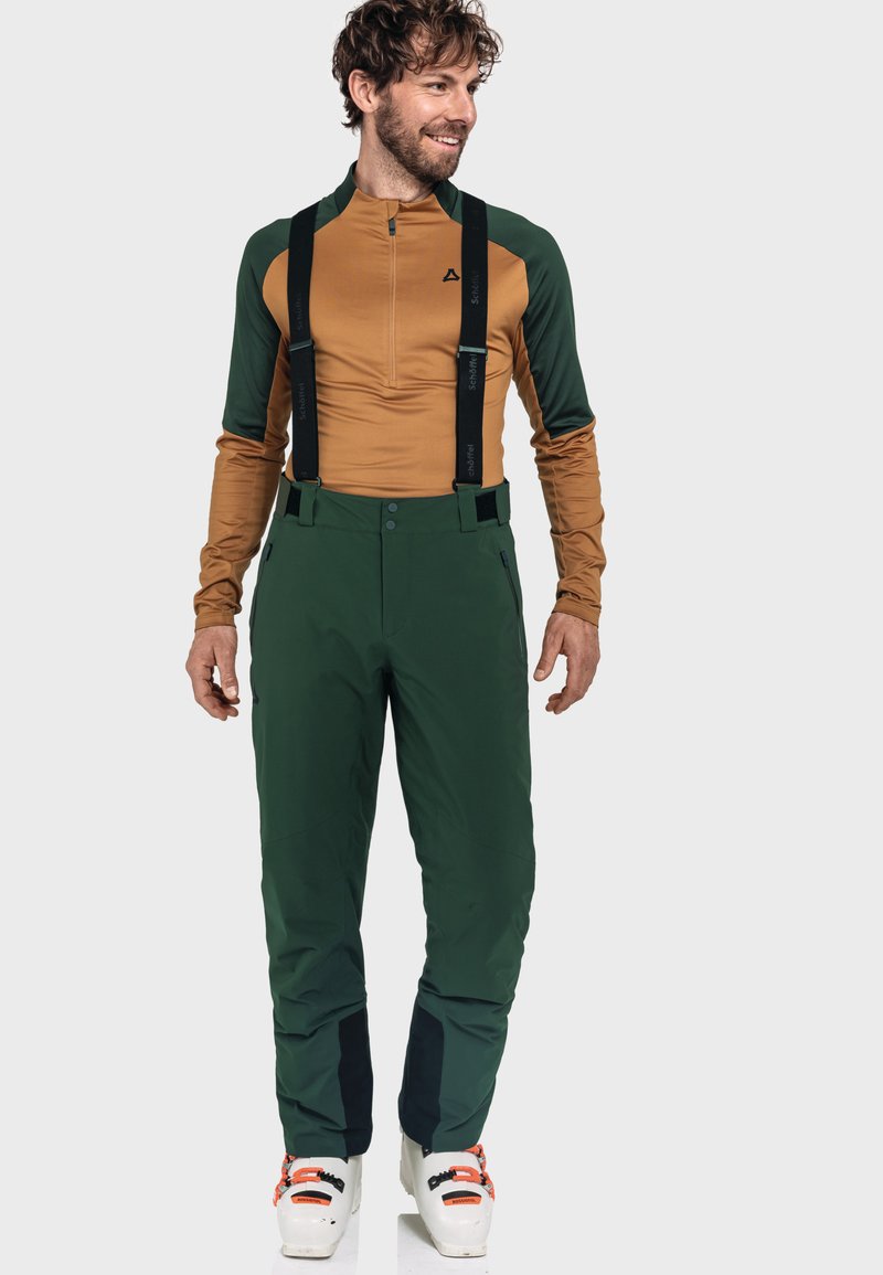 Mann in grünen Skihosen mit Trägern, braun-grünem Langarmshirt und weißen Skischuhen, der steht und seitlich schaut.