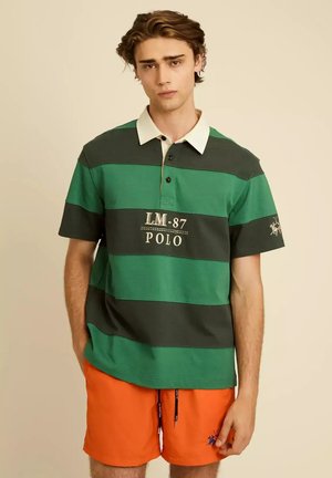 Joven vestido con polo de rayas verdes y negras con texto "LM-87 POLO" y pantalones cortos naranja brillante, de pie sobre un fondo beige.