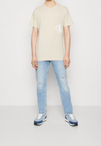 Camiseta de algodón beige con cuello redondo y logo blanco en el pecho, combinada con jeans claros rasgados y zapatillas con detalles azules.