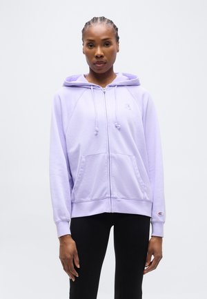 Γυναίκα με πλεξούδες, που φοράει ένα ανοιχτό μωβ φούτερ με φερμουάρ και μαύρο παντελόνι, στέκεται μπροστά σε ένα απλό λευκό φόντο.