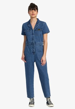 Denim jumpsuit with short sleeves, front pockets, and a fitted waist. Features a blue wash and tapered legs, paired with black sneakers.