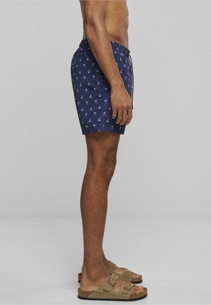 Egy férfi tengerészkék fürdőnadrágot visel fehér horgonydíszekkel és barnás csatos szandált, oldalról áll egy egyszerű szürke háttér előtt.