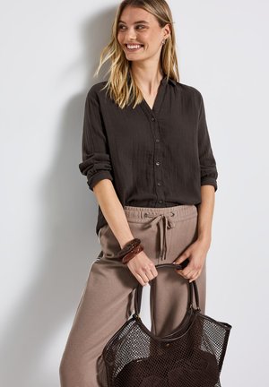 Mørkebrun skjorte med knapper og opbrettede ærmer, parret med lysebrune joggers. Modellen holder en stor mesh-totebag og har ambersfarvede armbånd.
