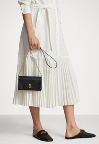 Borsa a mano nera in pelle con hardware dorato, abbinata a un vestito bianco plissettato e mule neri senza tacco. Il vestito presenta un cinturino in vita.