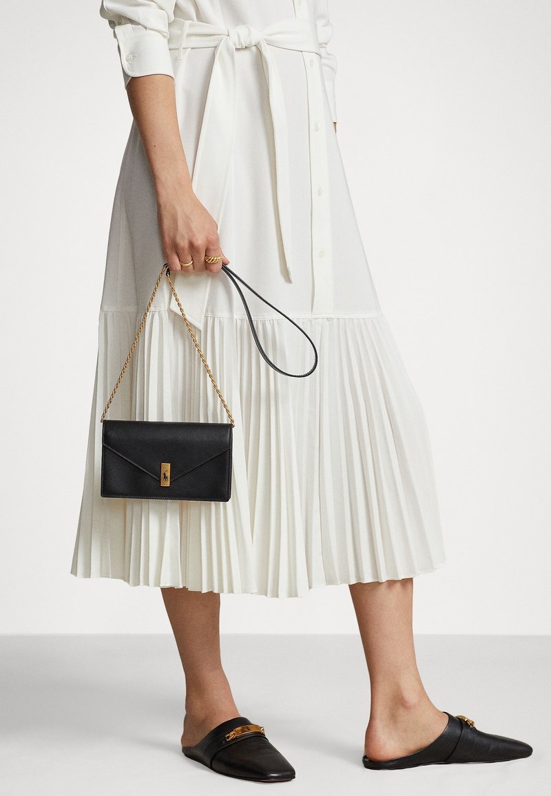 Borsa a mano nera in pelle con hardware dorato, abbinata a un vestito bianco plissettato e mule neri senza tacco. Il vestito presenta un cinturino in vita.