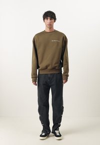 Oliven grønn genser med rund hals og brodert tekst, sammen med løstsittende svarte jeans med trykknapper og mørke sneakers.