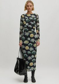 Langes schwarzes Kleid mit floralem Muster, das gelbe und grüne Rosen zeigt. Das Kleid hat lange Ärmel und ein drapiertes Design, kombiniert mit einer schwarzenTotetasche.