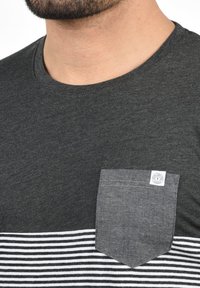 Camiseta gris de manga corta con un patrón de rayas en la parte inferior y un bolsillo oscuro con una etiqueta en el lado izquierdo. Textura suave.