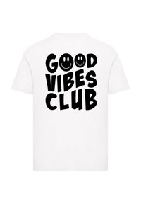 Bílá krátká tričko s krátkým rukávem, s výrazným černým textem: "KLUB DOBRÝCH VIBRACÍ" a dvěma ikonami usměvavých obličejů, hladká textilie.