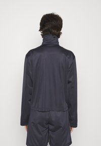 Mörk marinblå idrottsjacka med hög krage, gjord av ett mjukt och lätt material. Har långa ärmar och en avslappnad passform.