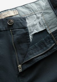 Pantalones slim-fit en tejido oscuro con cremallera, cierre de botón y forro a rayas en contraste. Detalle de costura visible en toda la prenda.