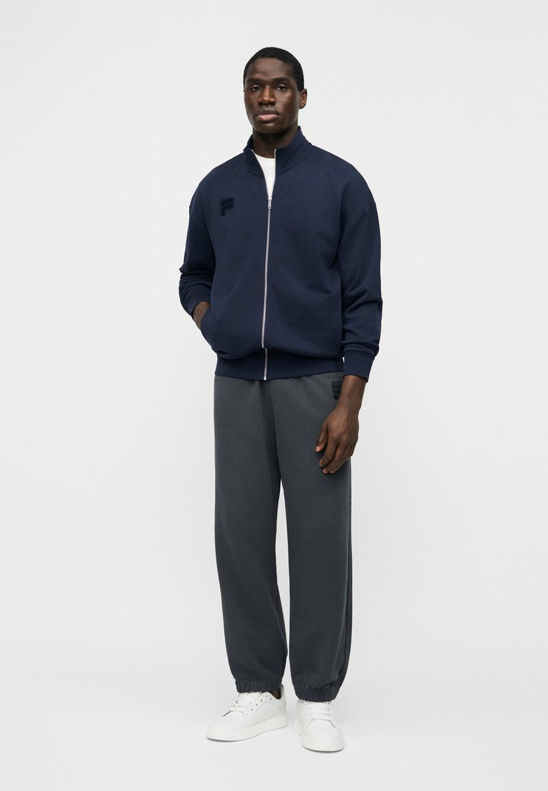 Homme debout, mains partiellement dans les poches, portant une veste zippée bleu marine, un pantalon de survêtement gris foncé et des baskets blanches, sur un fond blanc uni.