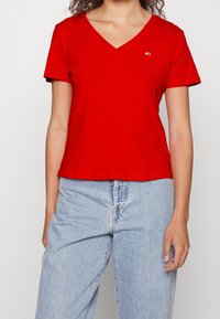 T-shirt in cotone rosso con scollatura a V e maniche corte. Presenta un piccolo logo sul petto. Abbinata a pantaloni di jeans leggeri blu.