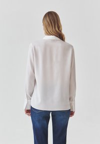 Bílá blůza s dlouhým rukávem a límcem, hladká textura a manžety na knoflíky, v kombinaci s tmavě modrými džíny. Zadní pohled ukazuje uvolněný střih.