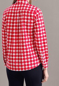 Czerwona koszula w białe kropki, z kołnierzykiem, długimi rękawami i mankietami zapinanymi na guziki. Materiał wydaje się gładki i lekki.