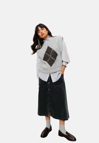 Un gilet en laine gris avec un motif en losanges noirs sur une chemise bleu clair. Une jupe en denim foncé, des chaussettes grises et des mocassins marron complètent la tenue.