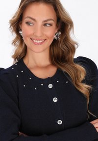Cardigan lavorato a maglia blu navy con maniche a sbuffo, caratterizzato da bottoni decorativi argentati e dettagli simili a cristalli attorno al colletto.