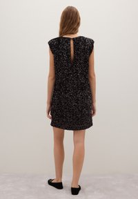 Femme portant une mini robe noire à sequins sans manches avec un détail en forme de trou ovale au dos, debout pieds nus sur un tapis clair contre un mur uni.