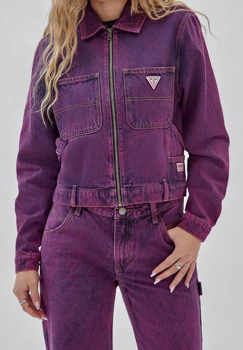 Paarse denimjas met een ritssluiting aan de voorkant, twee borstzakken en een klein driehoekig logo. De stof heeft een licht gestructureerde afwerking.