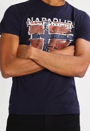 Henkilö, jolla on käsivarret ristissä ja joka pukeutunut tummansiniseen t-paitaan, jossa on kulunut Norjan lipun kuvio ja teksti "NAPAPIJRI".