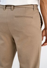 Calças castanho claro com textura suave, apresentando um bolso traseiro único e um design ajustado que contorna a cintura.