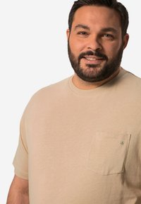 Béžové bavlněné tričko s krátkým rukávem a kulatým výstřihem, které má malou kapsu na levé straně hrudi se znakem. Hladká textura a ležérní střih.