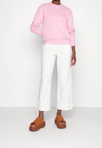 Lichtroze sweatshirt met de tekst "Hugs", gecombineerd met witte jeans met wijde pijpen en bruine slip-on platformschoenen. Soepele stof, ontspannen pasvorm.