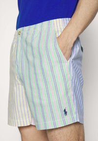 Bañador a rayas en verde, amarillo y azul. Cuenta con una banda elástica en la cintura, dos bolsillos laterales y un pequeño logotipo en el dobladillo.