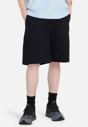 Czarne szorty o prostym kroju, boczne kieszenie oraz gładka bawełniana tkanina. Noszone z szarymi i czarnymi butami sportowymi oraz czarnymi skarpetami.