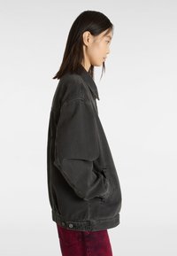 Zwarte denimjas met een klassieke kraag, relaxte pasvorm, zijzakken en knoopdetails op de taille, gedragen met rode jeans.