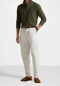 Olive green long-sleeve polo, white pleated trousers, brown leather belt, brown loafers, silver watch. Plain background.