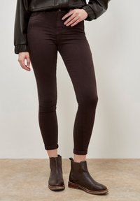 Jeans ajustados de color marrón oscuro con una textura suave, cintura alta y puños enrollados, combinados con unas botas de cuero negras hasta el tobillo con tacón bajo.