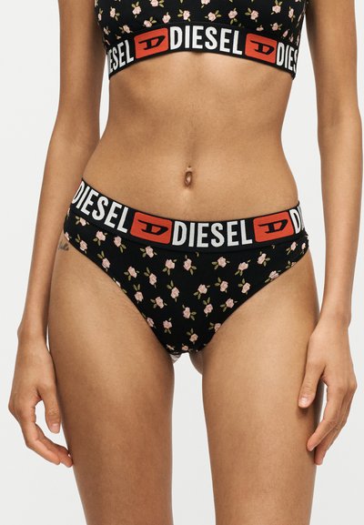 Femme portant des sous-vêtements noirs à motif floral et un soutien-gorge assorti avec le logo visible de la marque Diesel sur la ceinture élastique.