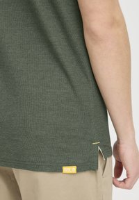 Grøn kortærmet t-shirt med struktureret stof, der har et gult mærke "ISOLID" på den nedre side og sidesplidser for bedre bevægelsesfrihed.