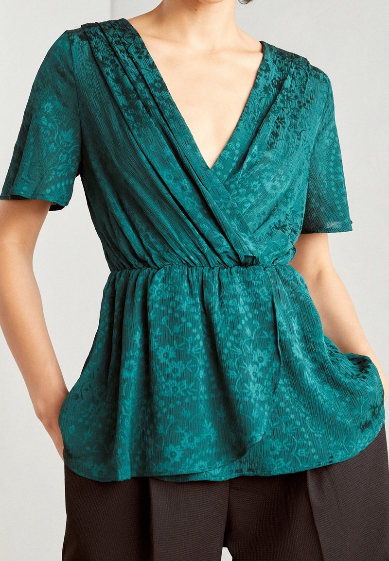 Blusa a stampa floreale turchese con scollatura a V profonda, maniche corte e vita arricciata. Realizzata in tessuto testurizzato, presenta dettagli plissettati.