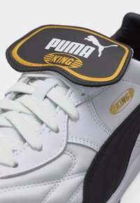 Valkoinen Puma King -tennari mustalla raidalla, valkoisilla nauhoilla ja mustalla kielenmerkillä, jossa on Puma-logo ja keltainen "King"-teksti.