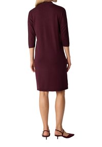 Donna che indossa un vestito viola scuro, a maniche lunghe e lunghezza al ginocchio, con scarpe a tacco basso e con cinturino alla caviglia abbinate, in piedi di spalle con le mani lungo i fianchi.