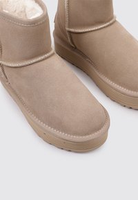 Botines de ante beige con suave forro de pelo, suelas de goma planas y costuras decorativas a lo largo de las costuras. Diseño de punta redondeada.